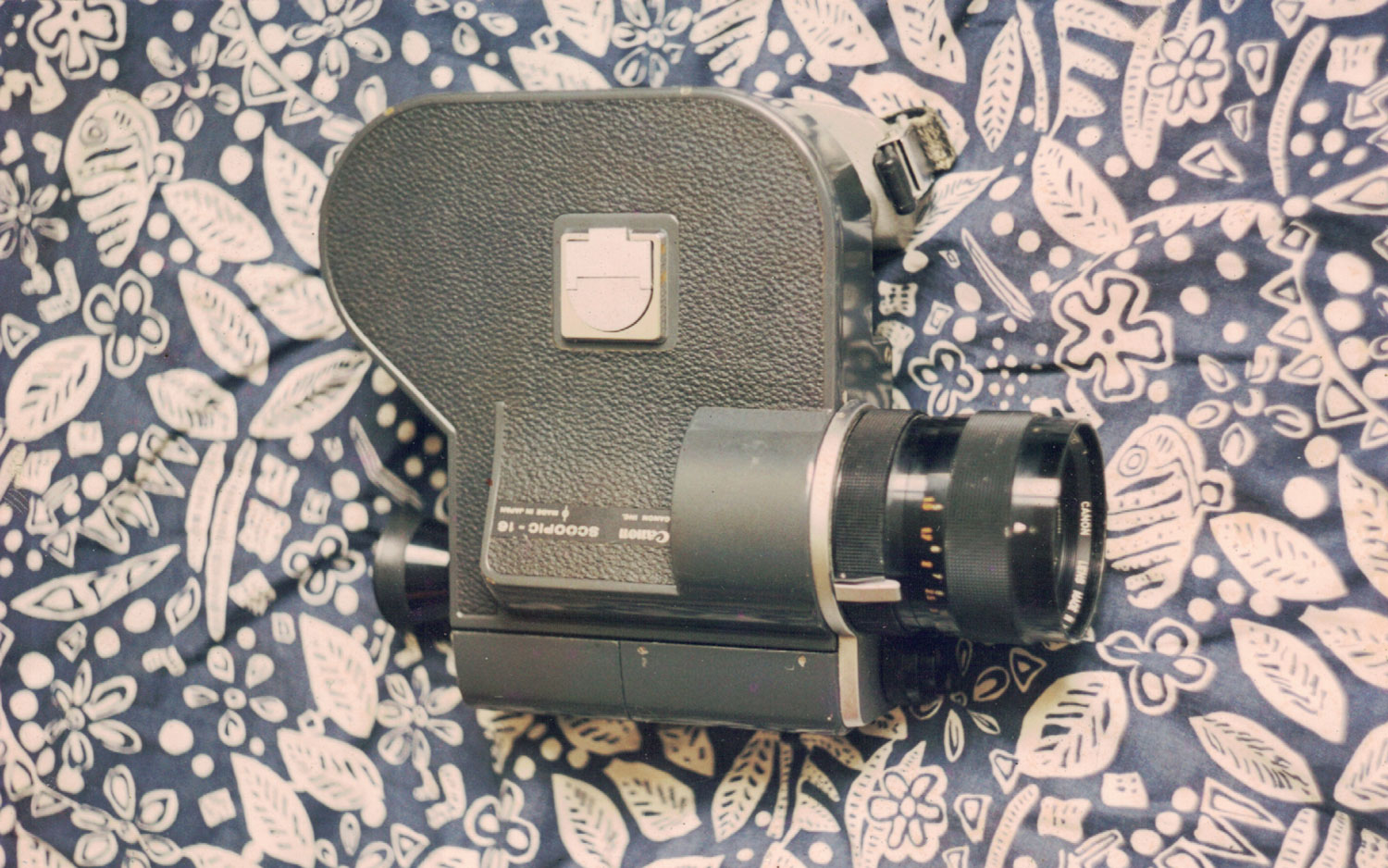 Câmera Canon Scoopic 16mm sobre tecido estampado nas cores azul e bege e tampa de lente ao seu lado. Fotógrafo(a) não identificado(a). 