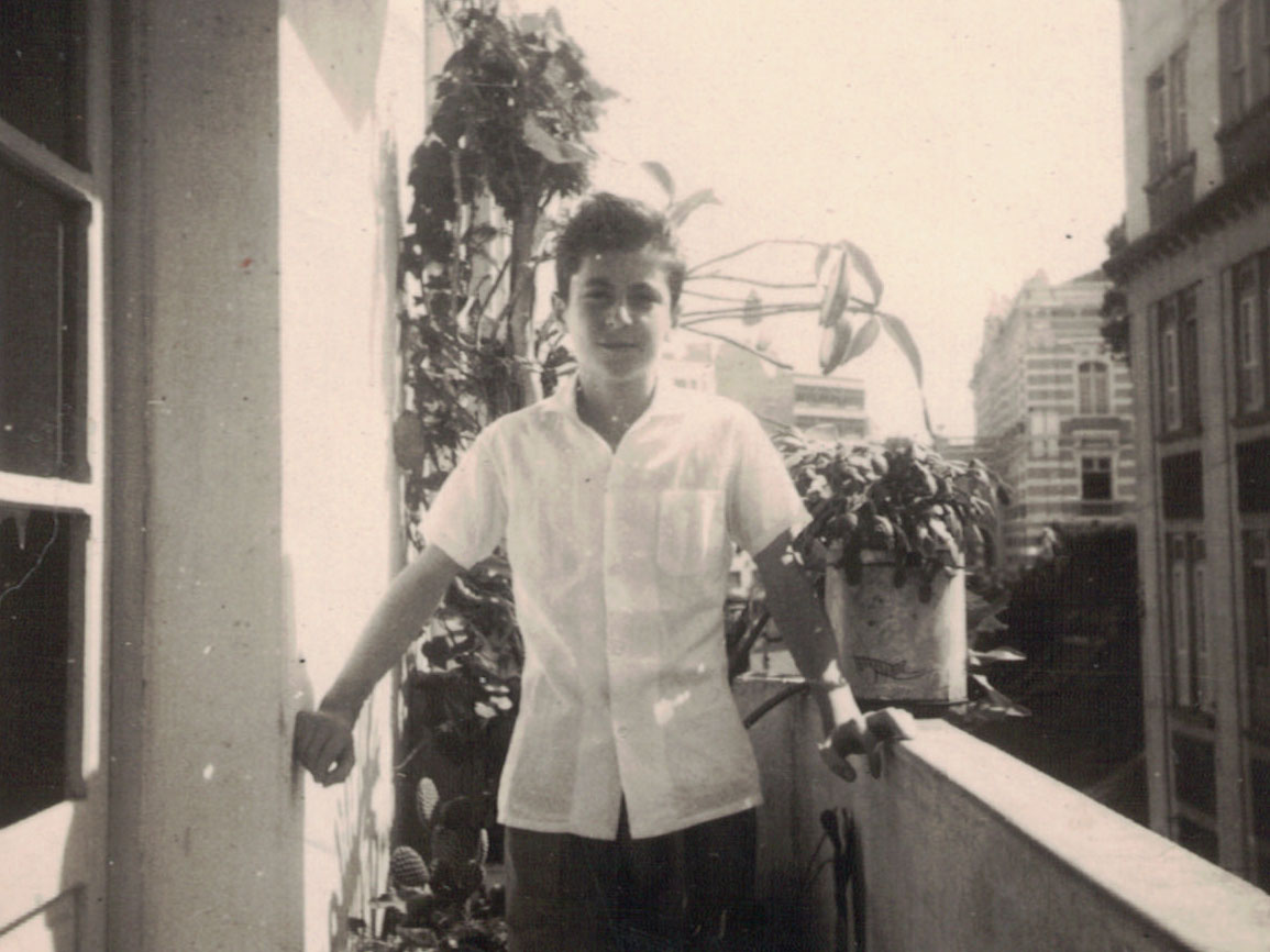 Ramon Alvarado quando criança, vestindo camisa social de mangas curtas, na varanda da casa de seus pais em Vitória (ES), com plantas ao seu lado.  Prédios ao fundo, entre eles o Palácio Anchieta.
